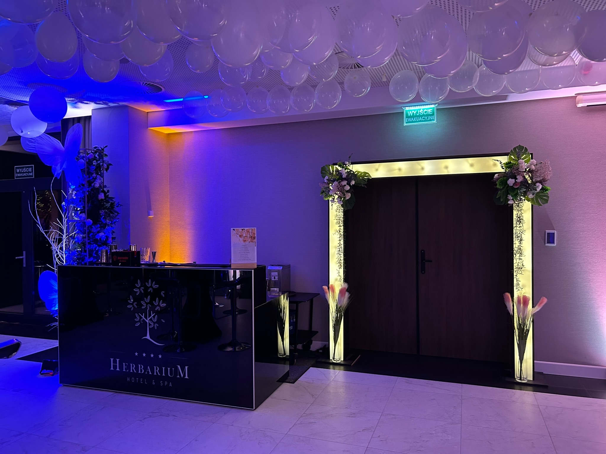 Eleganckie wejście do sali z czarnym barem z logo Herbarium Hotel & SPA, dekoracjami kwiatowymi, oświetlonymi kolumnami i białymi balonami pod sufitem.