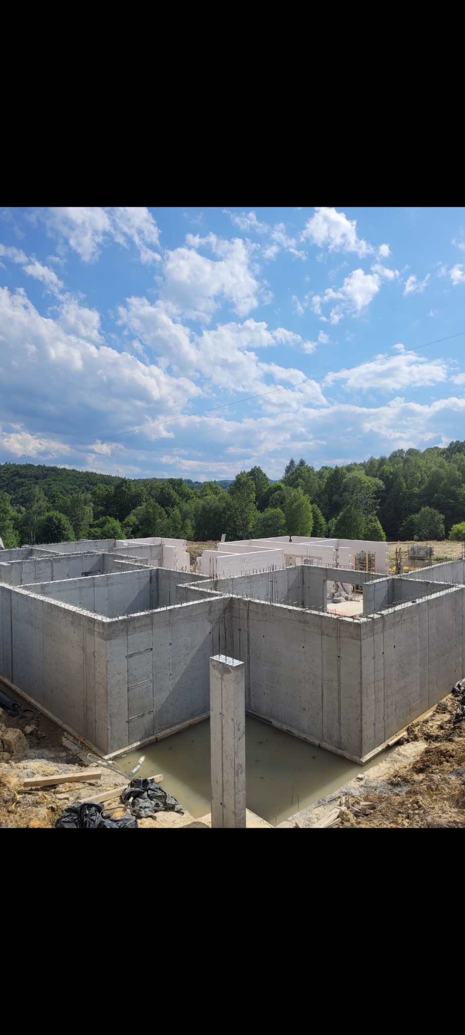 Wylane betonowe fundamenty domu z widocznym zbrojeniem, częściowo wypełnione wodą, na tle lasu i błękitnego nieba z chmurami.