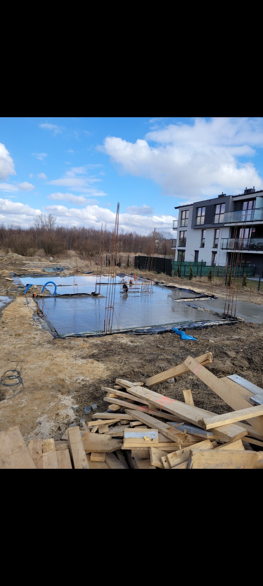 Zalany fundament z wystającymi prętami zbrojeniowymi, widoczne deski szalunkowe i nowy budynek w tle.