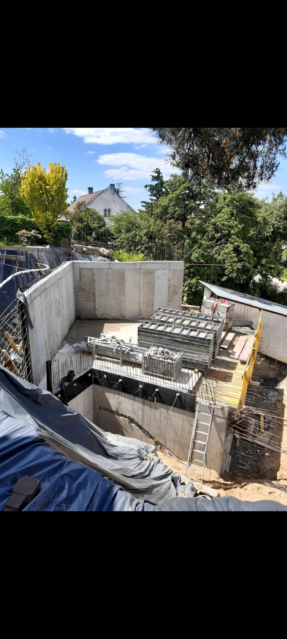 Wykonane betonowe ściany fundamentowe z wystającymi prętami zbrojeniowymi, zabezpieczone folią hydroizolacyjną, przygotowane do dalszych prac budowlanych na działce z widokiem na dom i zieleń.