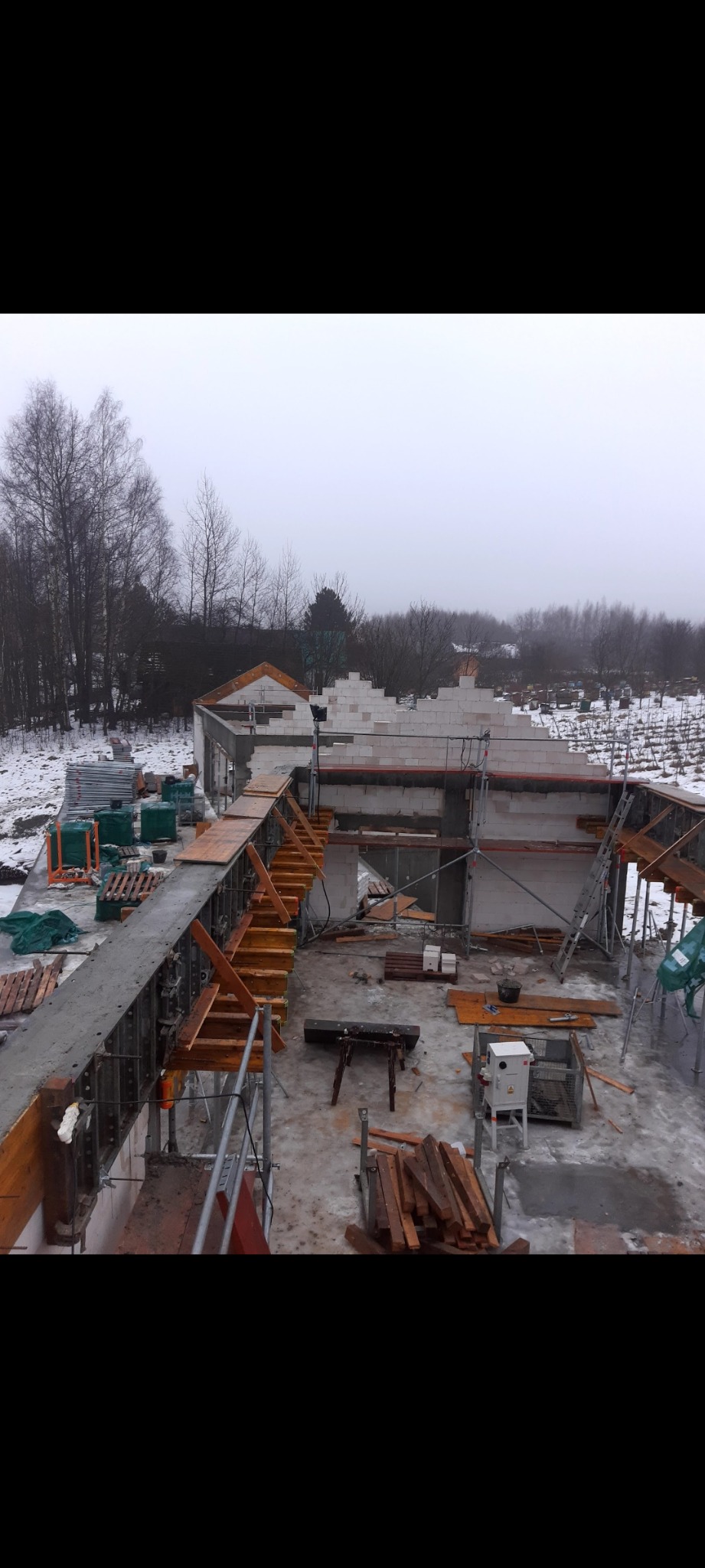 Budowa domu w stanie surowym otwartym w zimowej scenerii, widoczne fundamenty, ściany z bloczków i szalunki, w tle zalesiony teren pokryty śniegiem.