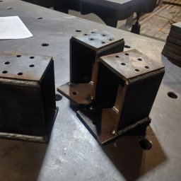 Ekoauto - Trzy metalowe, spawane konstrukcje w kształcie prostopadłościanów z otworami, leżące na metalowym stole spawalniczym. Widoczne ślady spawania i obróbki metalu.