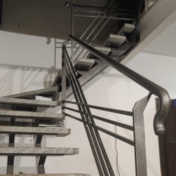 Ekoauto - Nowoczesne schody metalowe z poręczą o zaokrąglonym zakończeniu, stopnie wykończone wzorem lastryko, widok z dołu na klatkę schodową.