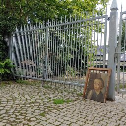 Ekoauto - Otwarta metalowa brama wjazdowa z ostrymi zakończeniami prętów, na brukowanym podjeździe, z obrazem opartym o słupek bramy.