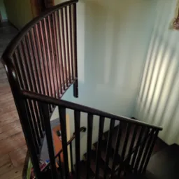 Spiralne schody wewnątrz domu z metalową balustradą w kolorze ciemnej miedzi, widok z góry na drewniane stopnie i podłogę, z obrazem w złotej ramie na ścianie.