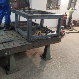 Ekoauto - Metalowa konstrukcja spawana, z użyciem zacisków, umieszczona na stole spawalniczym z rastrem otworów, w tle okna warsztatowe i sprzęt spawalniczy.