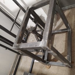 Ekoauto - Surowa, metalowa konstrukcja ramy schodów na etapie spawania, widoczne ślady po spawaniu, ustawiona w warsztacie na betonowej posadzce, z widocznymi elementami rusztowania w tle.