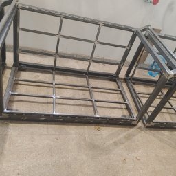 Ekoauto - Metalowe ramy konstrukcyjne z profili kwadratowych, spawane, ułożone na podłodze w warsztacie, widoczne spawy i niedoskonałości obróbki.