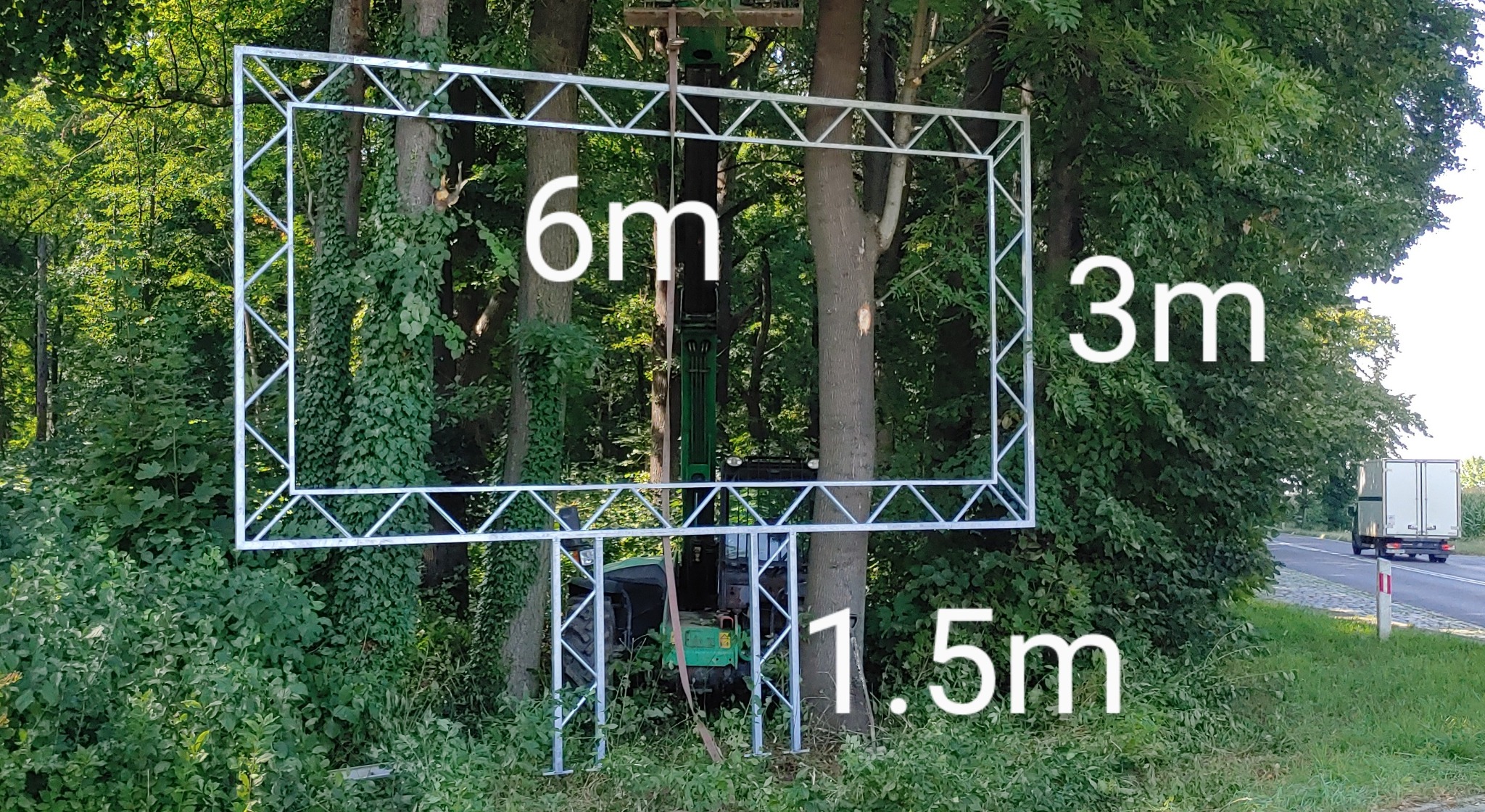 Stalowa rama o wymiarach 6x3 metry i wysokości 1.5 metra, konstrukcja kratowa, ustawiona w zaroślach z widocznym traktorem w tle, prawdopodobnie przygotowana do montażu reklamy.