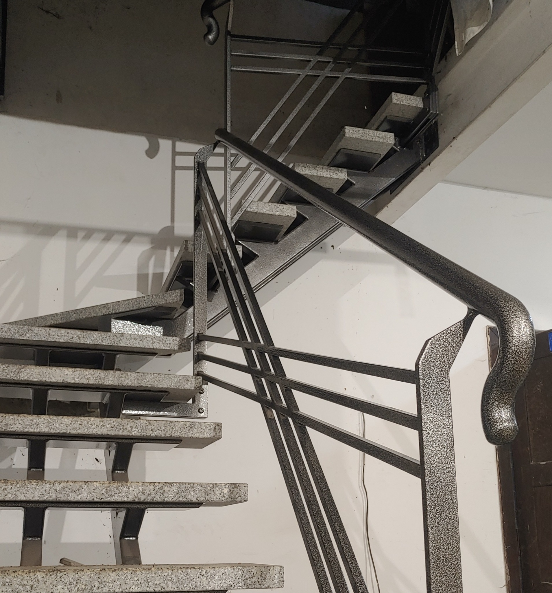 Nowoczesne schody metalowe z poręczą o zaokrąglonym zakończeniu, stopnie wykończone wzorem lastryko, widok z dołu na klatkę schodową.