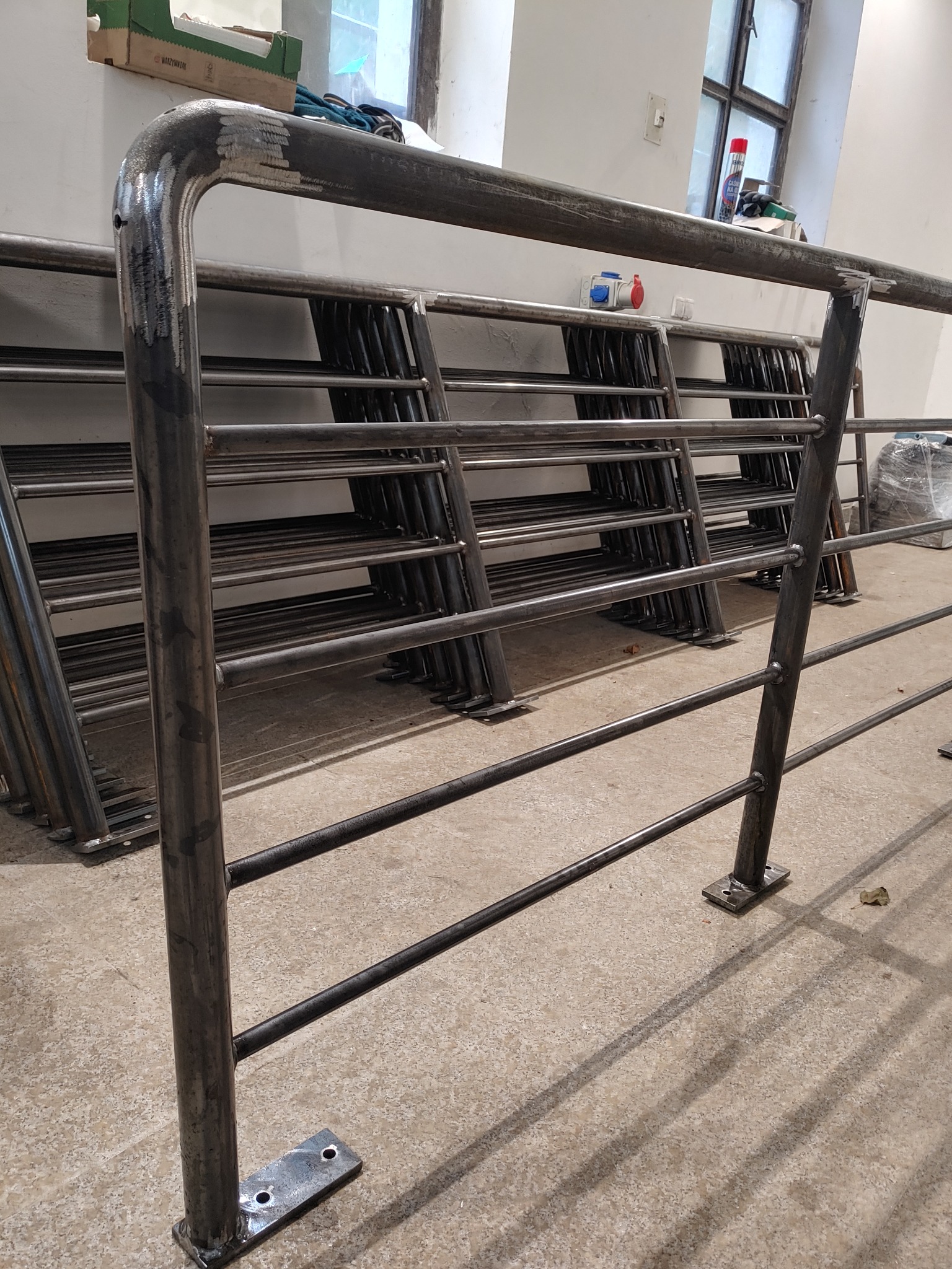 Stos metalowych balustrad schodowych, częściowo wyszlifowanych, w warsztacie, gotowych do montażu, z widocznymi spawami i elementami mocującymi.