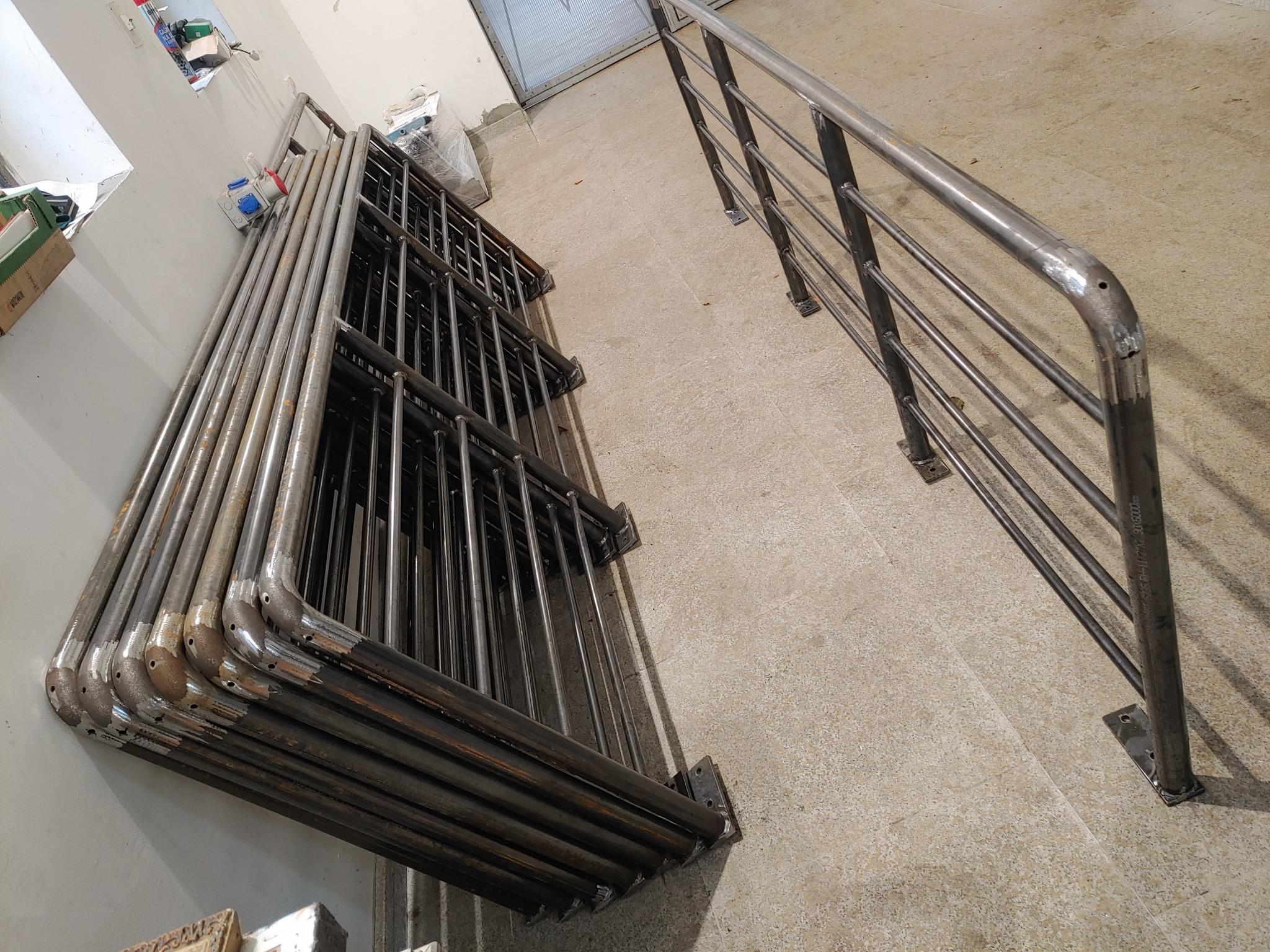 Stos metalowych balustrad schodowych, częściowo obrobionych, opartych o ścianę, obok zamontowana, pojedyncza metalowa barierka na betonowej posadzce.