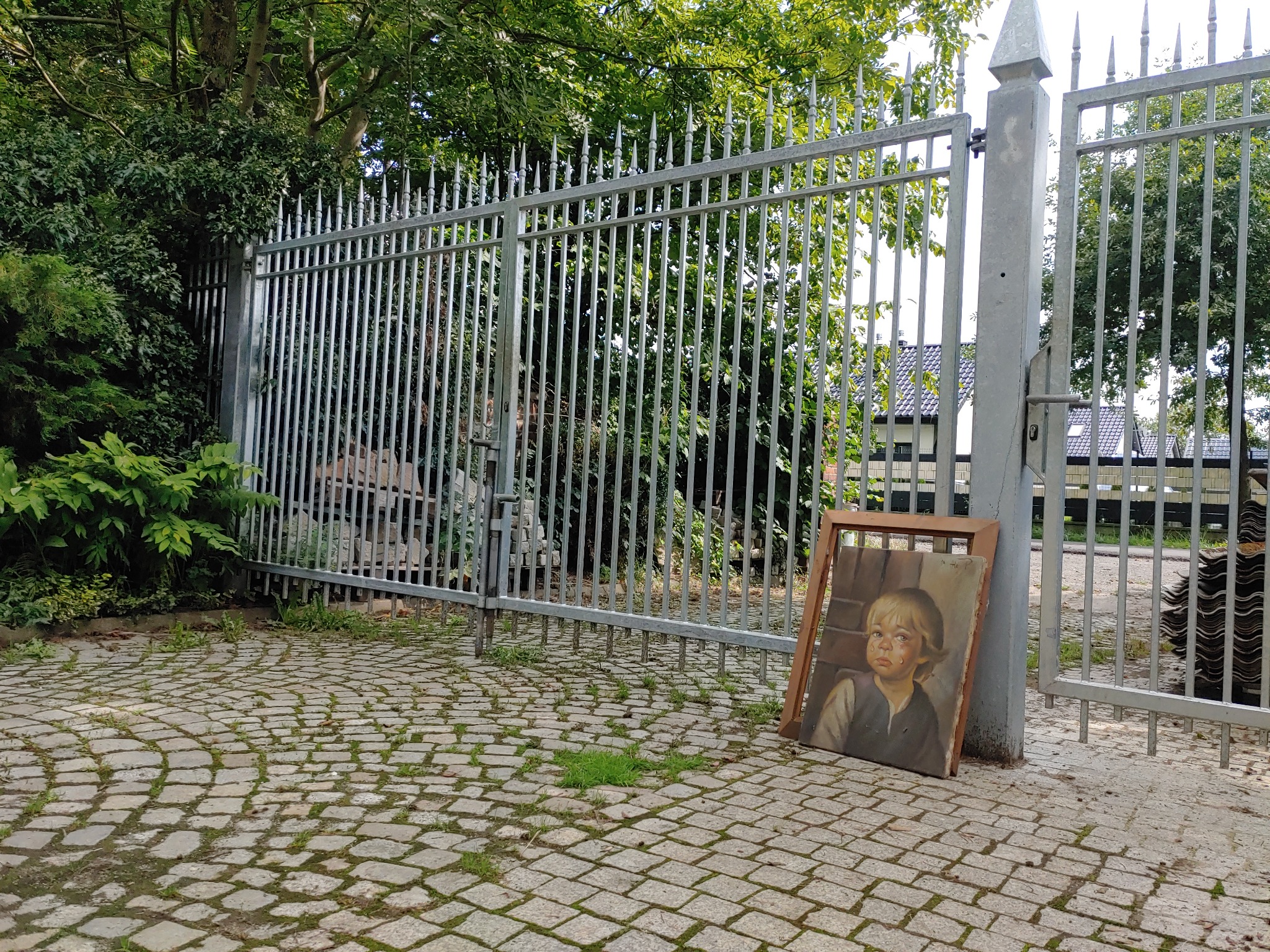 Otwarta metalowa brama wjazdowa z ostrymi zakończeniami prętów, na brukowanym podjeździe, z obrazem opartym o słupek bramy.