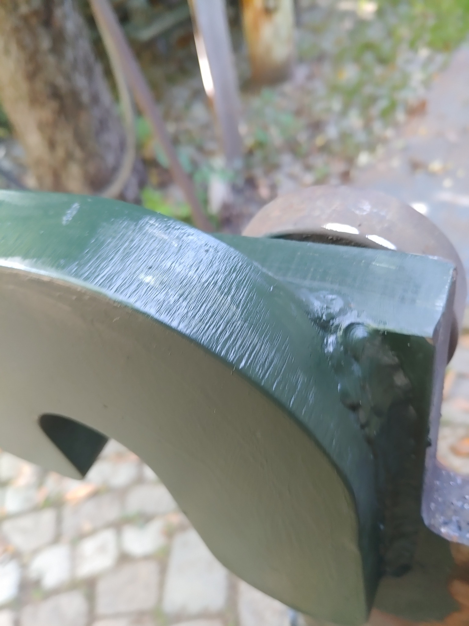 Fragment pomalowanego na zielono elementu metalowego z wyciętym wzorem, mocowanego w imadle, w tle widoczne drzewa i ścieżka.