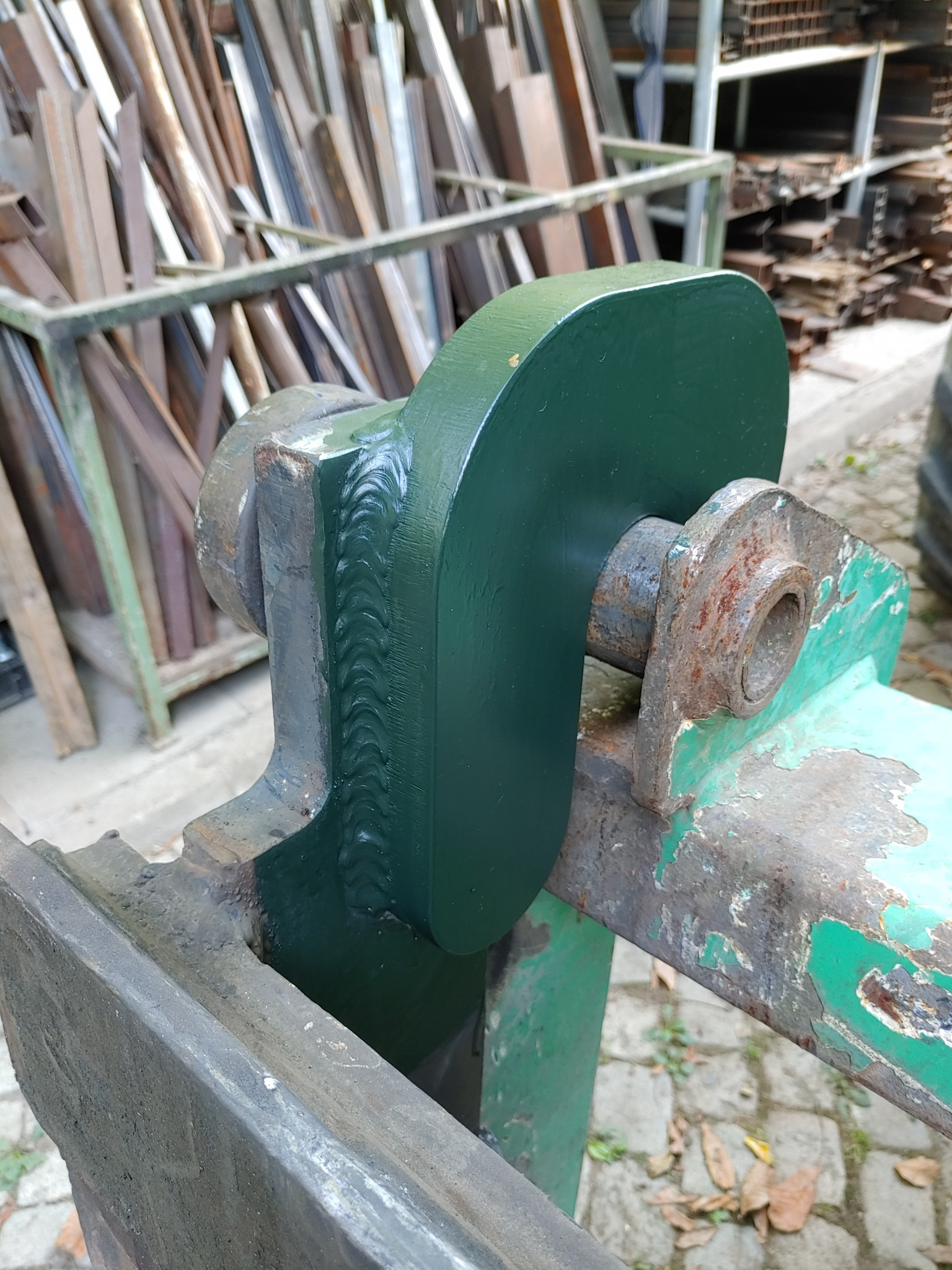 Fragment konstrukcji stalowej z widocznymi spawami, pomalowanej na zielono, z elementami rdzawymi. W tle składowane profile metalowe.