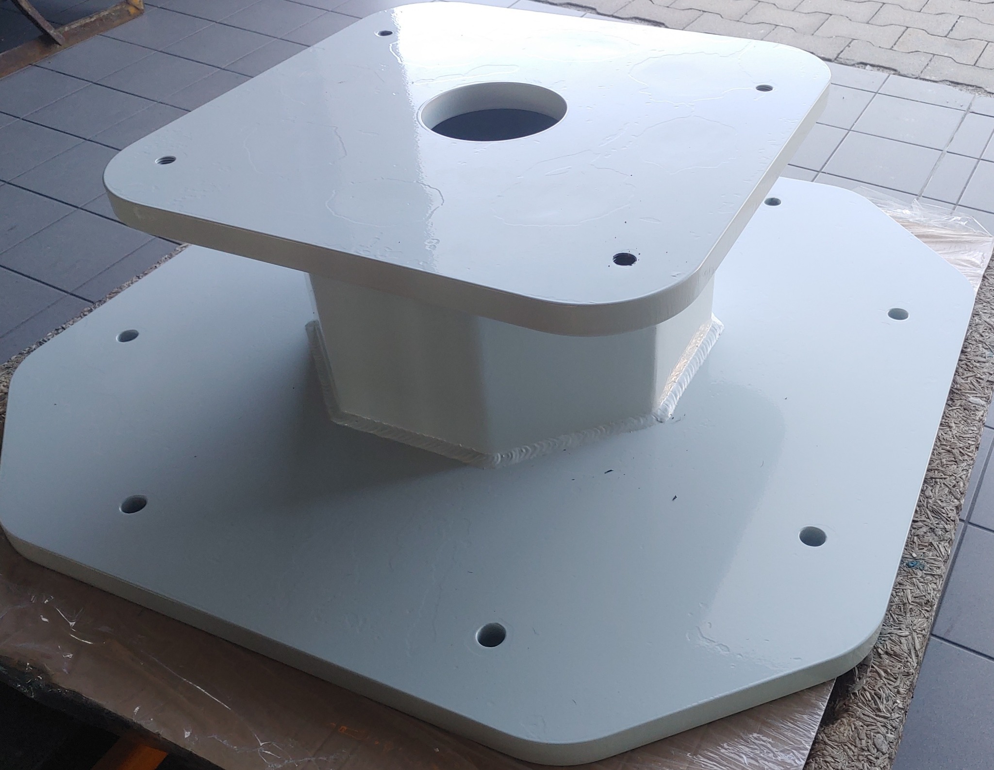 Biała, pomalowana proszkowo, stalowa podstawa konstrukcyjna z otworem centralnym i otworami montażowymi, widoczne spawy.