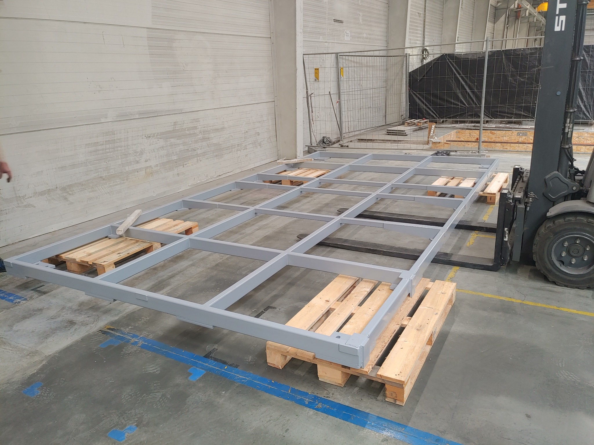 Szara, stalowa rama konstrukcyjna podzielona na kwadraty, umieszczona na drewnianych paletach, transportowana w hali produkcyjnej za pomocą wózka widłowego.