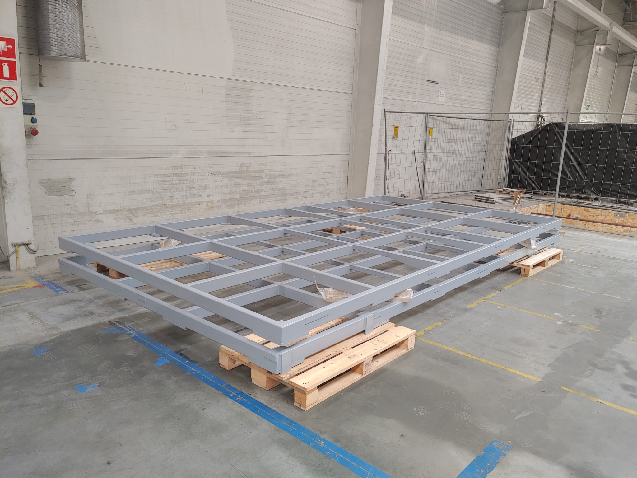 Gotowa, pomalowana na szaro, stalowa rama konstrukcji hali, ułożona na drewnianych paletach w hali produkcyjnej, widoczne ślady użytkowania na podłodze.