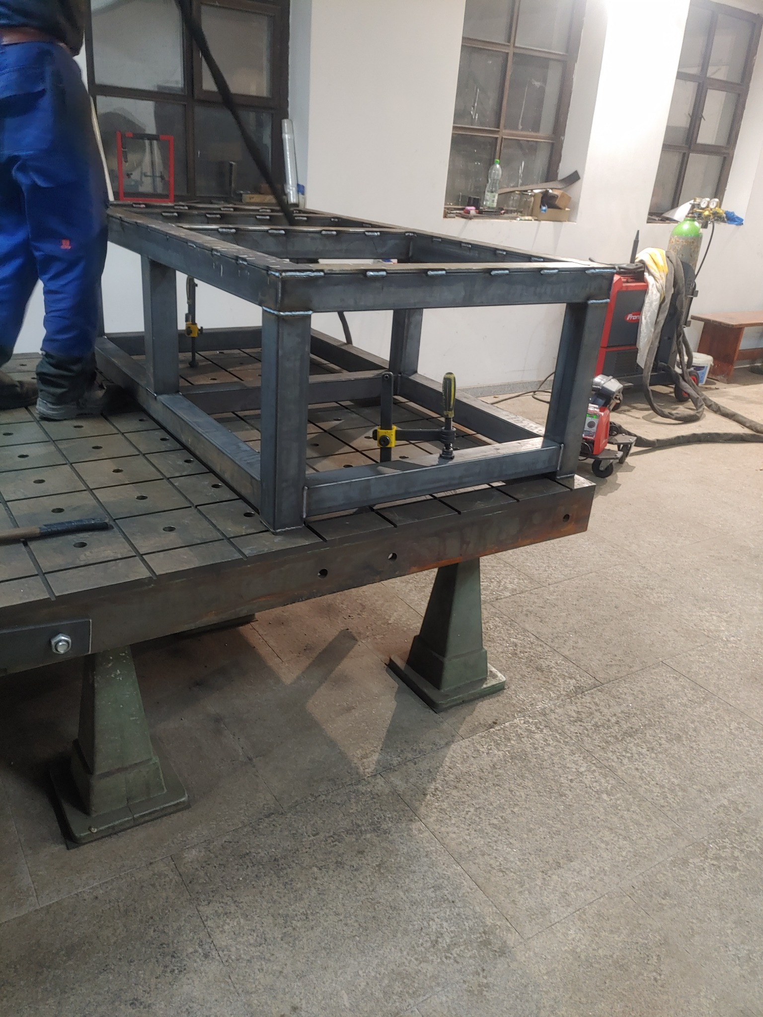 Metalowa konstrukcja spawana, z użyciem zacisków, umieszczona na stole spawalniczym z rastrem otworów, w tle okna warsztatowe i sprzęt spawalniczy.