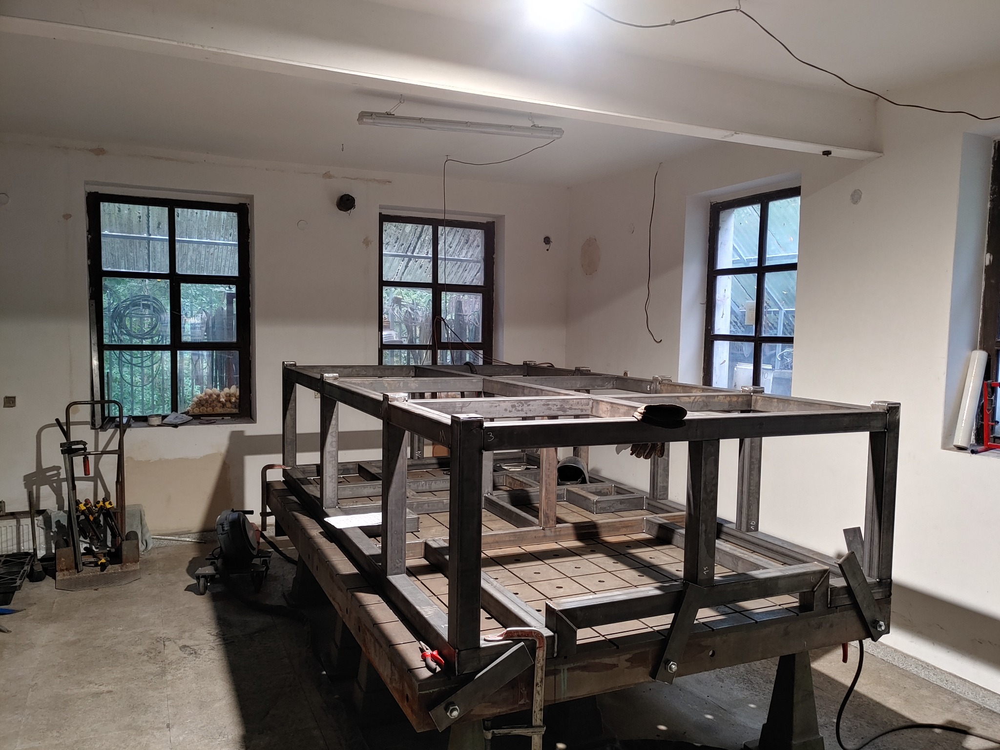 Metalowa konstrukcja szkieletowa na stole warsztatowym w jasnym pomieszczeniu, widoczne narzędzia i elementy montażowe, surowe wykończenie, trzy okna w tle.