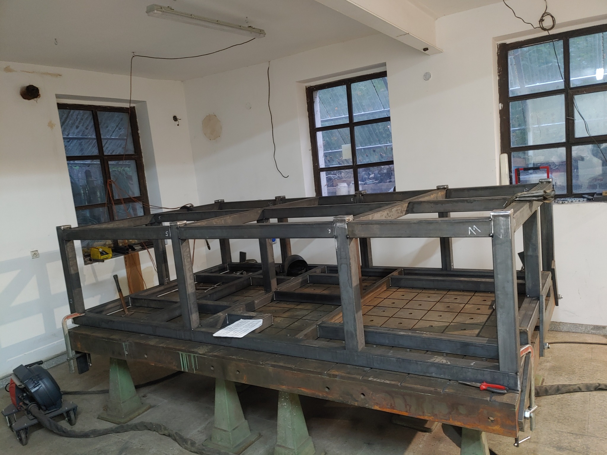 Konstrukcja stalowa w trakcie spawania, widoczne okna w tle, leżące narzędzia i dokumentacja.