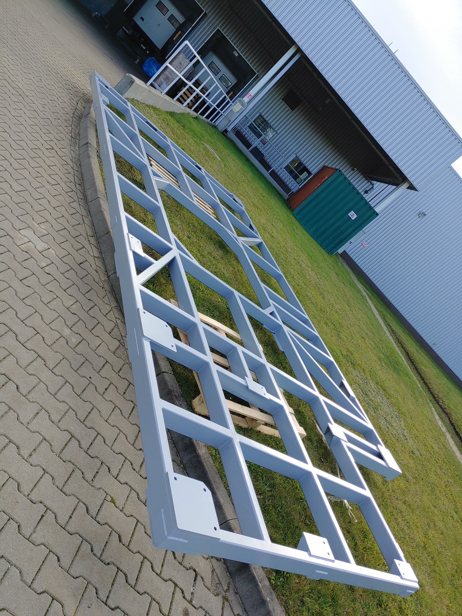 Szara, stalowa rama konstrukcyjna z profili, leżąca na palecie na trawie, przed budynkiem przemysłowym z rampami załadunkowymi.