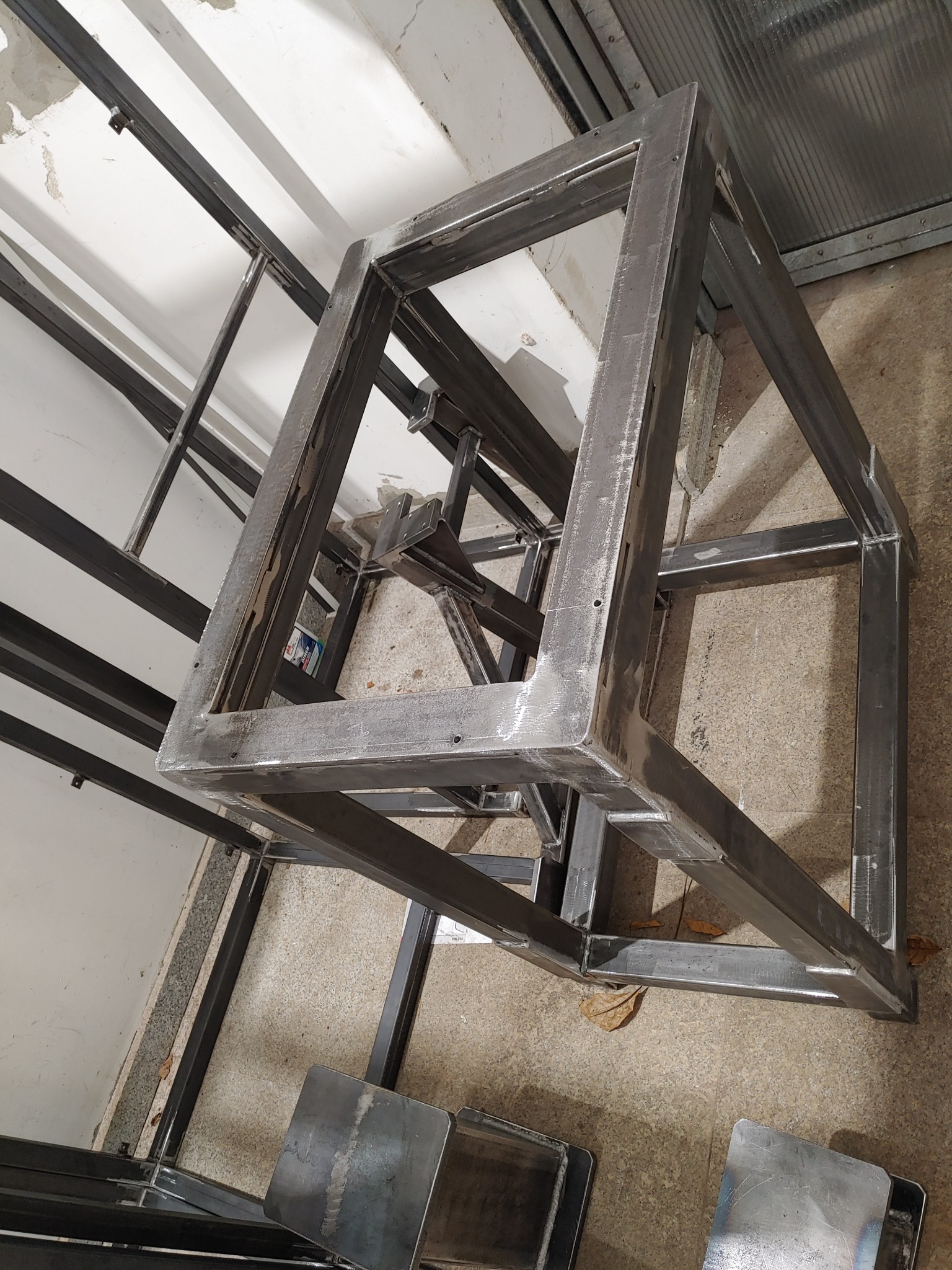 Surowa, metalowa konstrukcja ramy schodów na etapie spawania, widoczne ślady po spawaniu, ustawiona w warsztacie na betonowej posadzce, z widocznymi elementami rusztowania w tle.