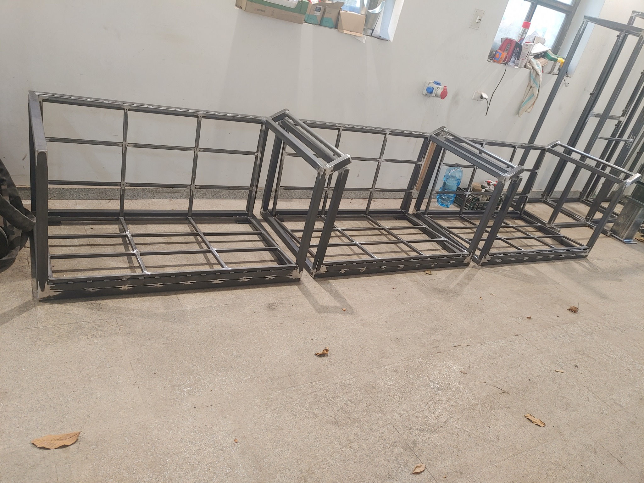 Częściowo zmontowane, spawane ramy z profili stalowych, prawdopodobnie do budowy konstrukcji, ustawione w warsztacie na betonowej posadzce, widoczne spawy i niedoskonałości, w tle okno i narzędzia.