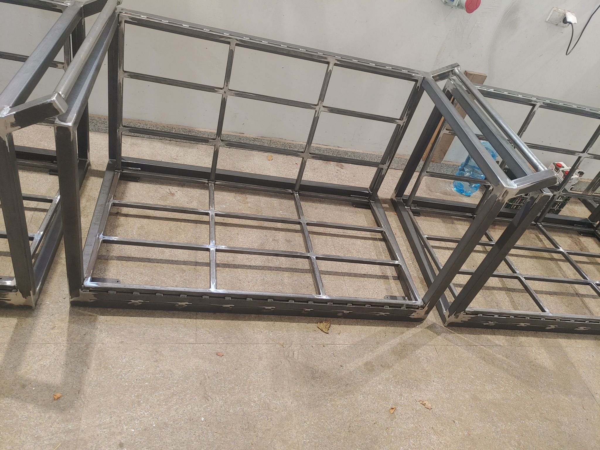 Metalowe ramy konstrukcyjne z profili kwadratowych, spawane, ułożone na podłodze w warsztacie, widoczne spawy i niedoskonałości obróbki.