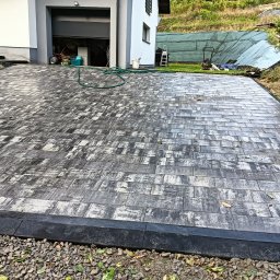 KOSTKA BRUKOWA I GRANITOWA - Ułożona kostka brukowa o nieregularnym wzorze szaro-białych prostokątów, obrzeża z czarnego betonu, w tle dom z otwartym garażem i sprzętem budowlanym.