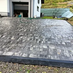 Ułożona kostka brukowa o nieregularnym wzorze szaro-białych prostokątów, obrzeża z czarnego betonu, w tle dom z otwartym garażem i sprzętem budowlanym.