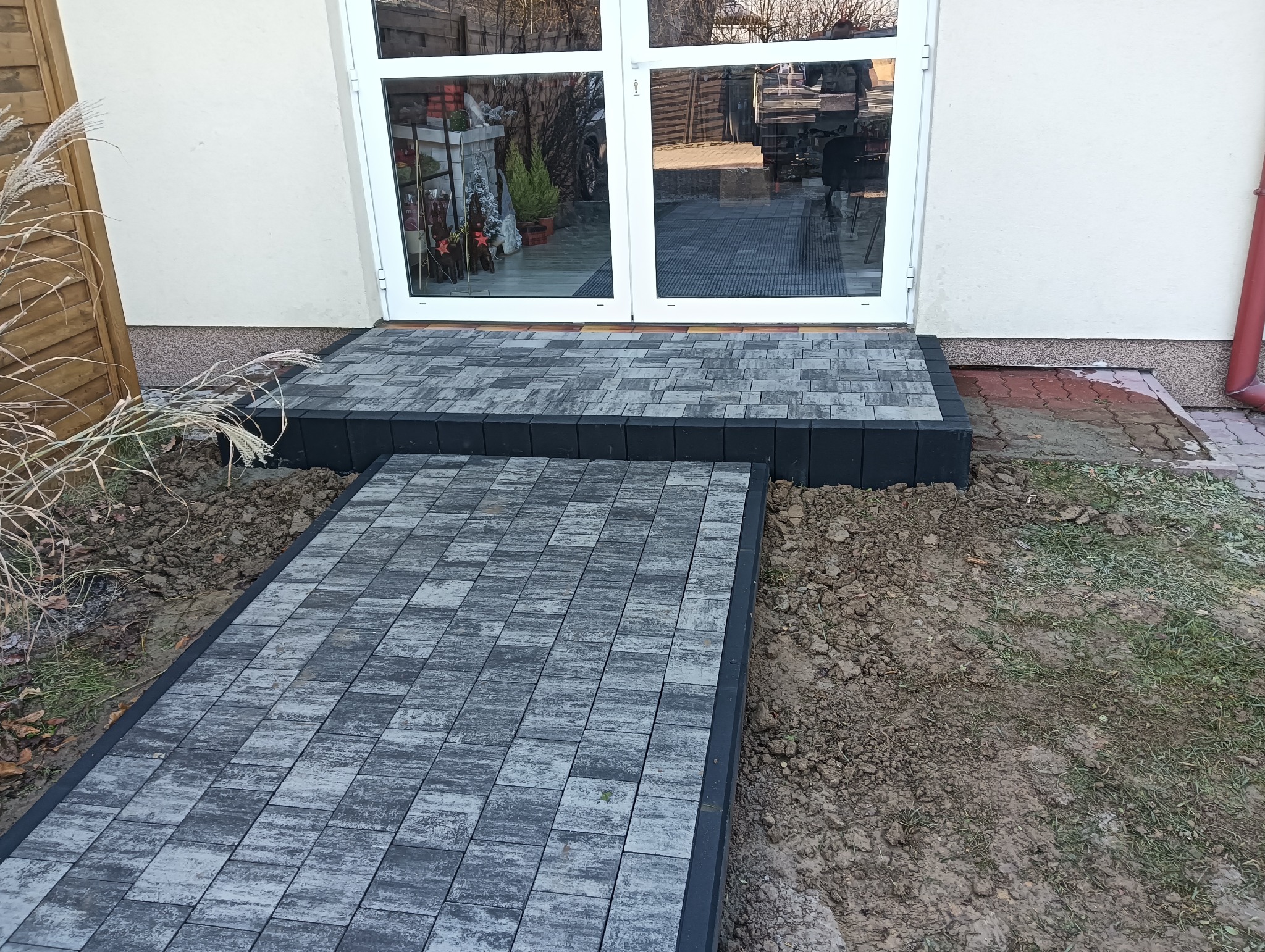 Ułożona kostka brukowa prowadząca do wejścia z białymi drzwiami, widoczne obrzeża i świeżo obsypana ziemia.