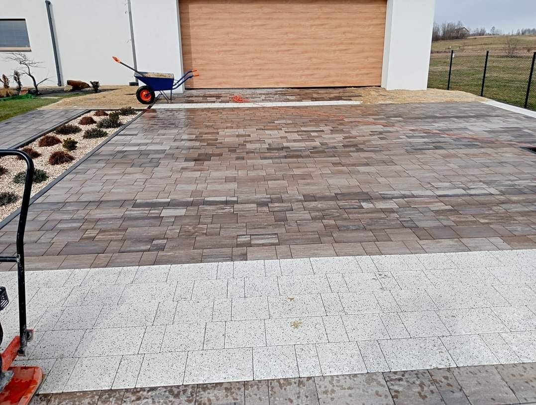 Ułożona kostka brukowa w odcieniach brązu i szarości z białym obramowaniem, widoczna taczka, zagęszczarka i fragment domu z bramą garażową.