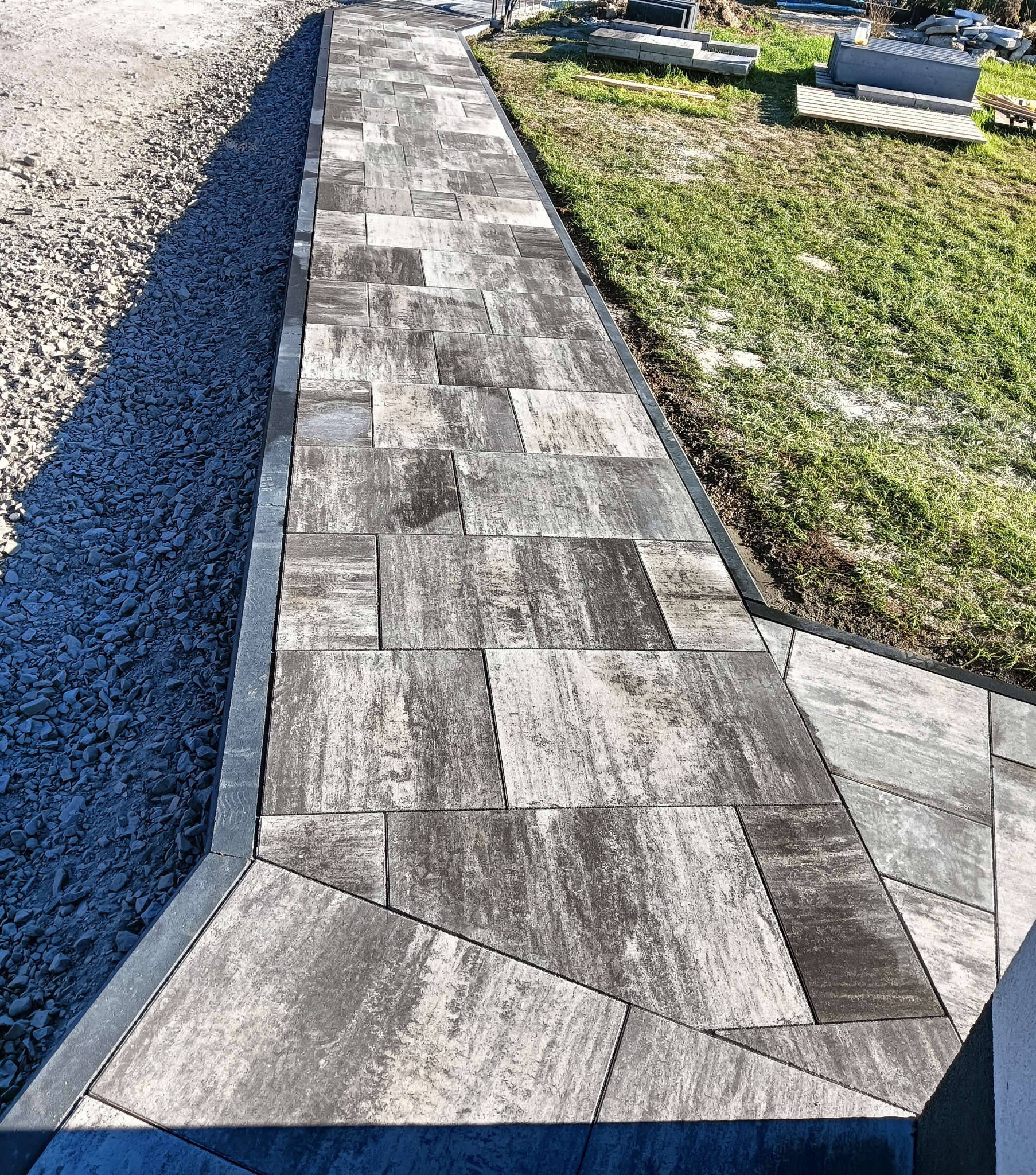 Ułożona ścieżka z szarych, nieregularnych płyt betonowych, obrzeża z metalową listwą, obok wysypany żwir i trawa, w tle stos płyt.