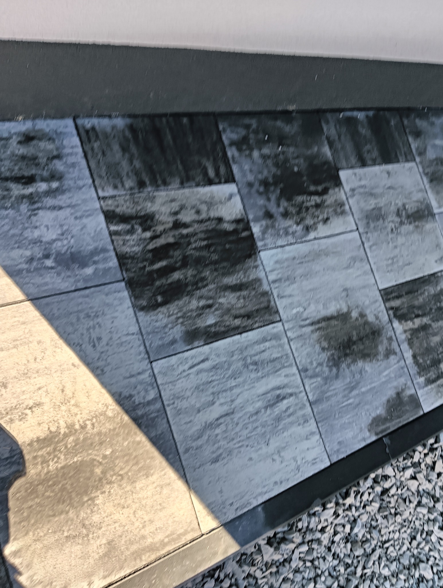 Ułożona, szara kostka brukowa z widoczną fakturą, obramowana czarnym obrzeżem, z fragmentem jasnego elementu architektonicznego w lewym dolnym rogu i drobnym żwirem po prawej stronie.