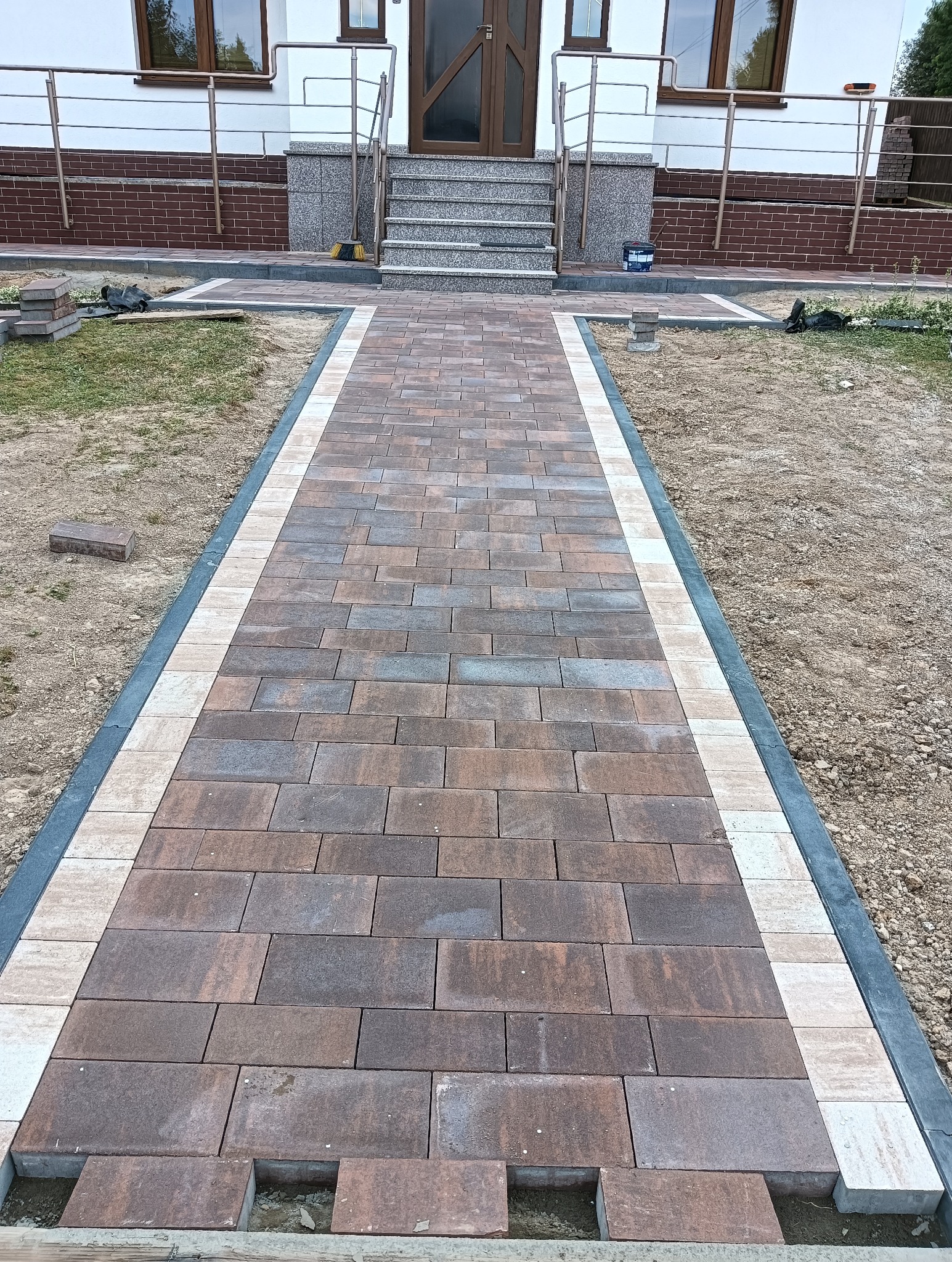 Ścieżka z kostki brukowej w odcieniach brązu i szarości prowadząca do wejścia domu z kamiennymi schodami, obrzeża z jasnej kostki, widoczne niedokończone fragmenty przy ziemi.