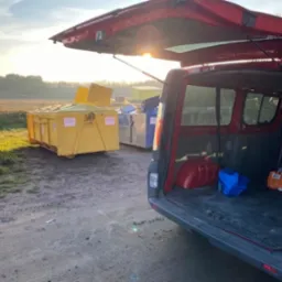 Otwarty bagażnik czerwonego samochodu dostawczego na tle żółtego kontenera na gruz z otwartą klapą i innych kontenerów w oddali, w porannym świetle.