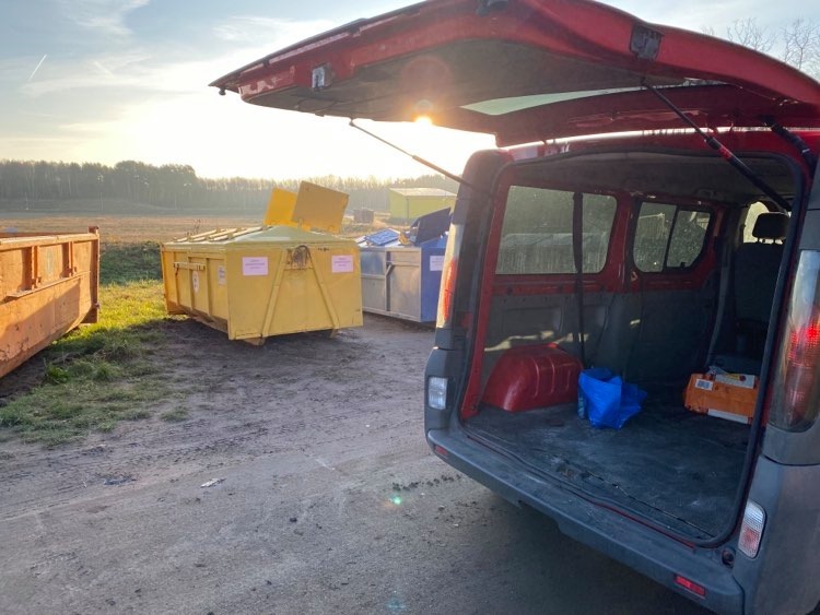 Otwarty bagażnik czerwonego samochodu dostawczego na tle żółtego kontenera na gruz z otwartą klapą i innych kontenerów w oddali, w porannym świetle.