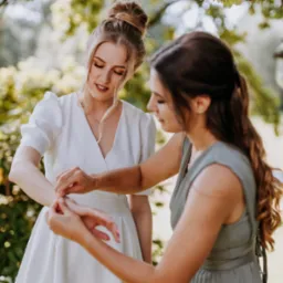 Panna młoda w białej sukni, której druhna zapina bransoletkę na nadgarstku w plenerze.