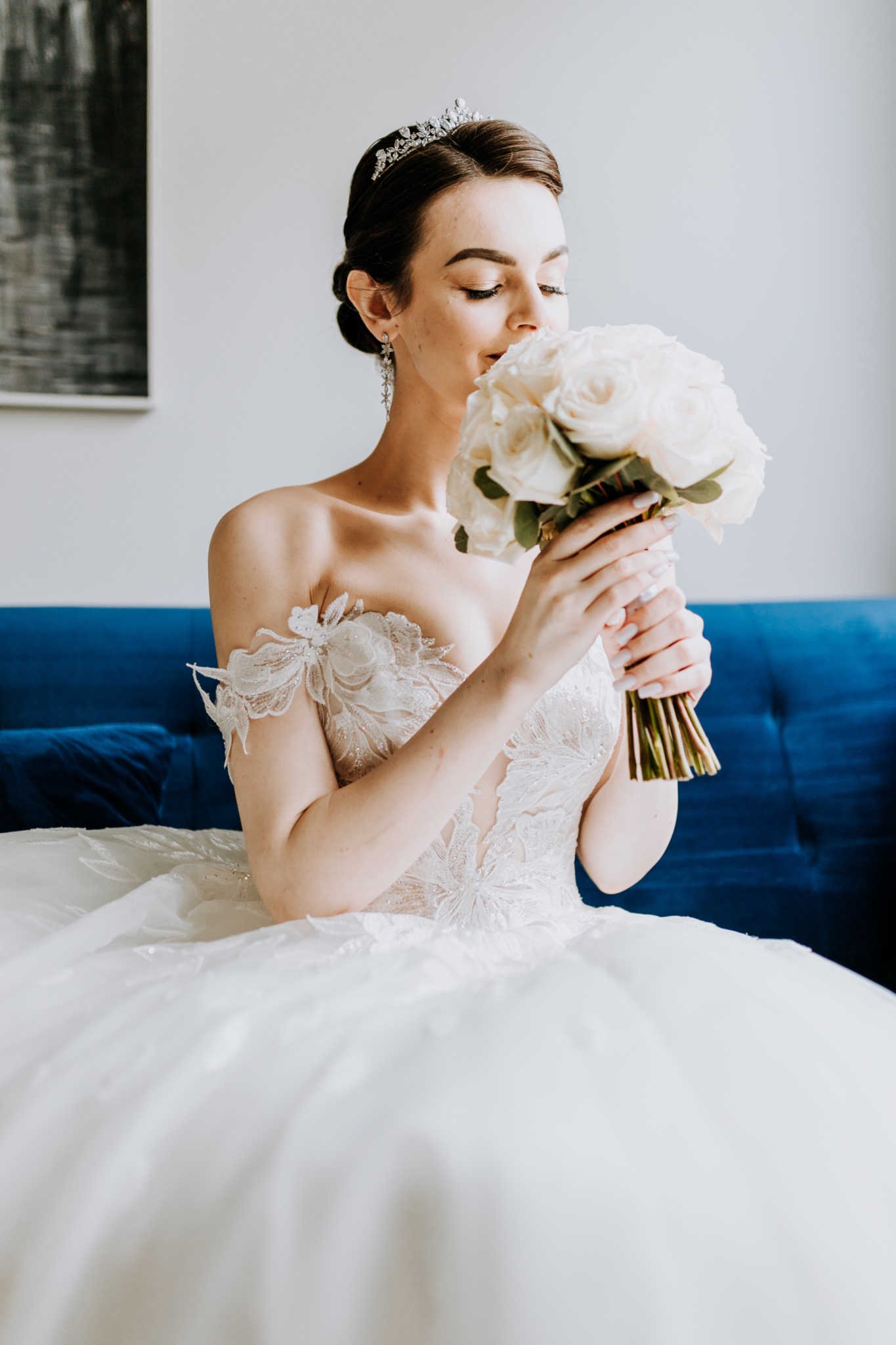 Panna młoda w koronie i sukni z koronkowymi aplikacjami, wącha bukiet białych róż na tle niebieskiej sofy.