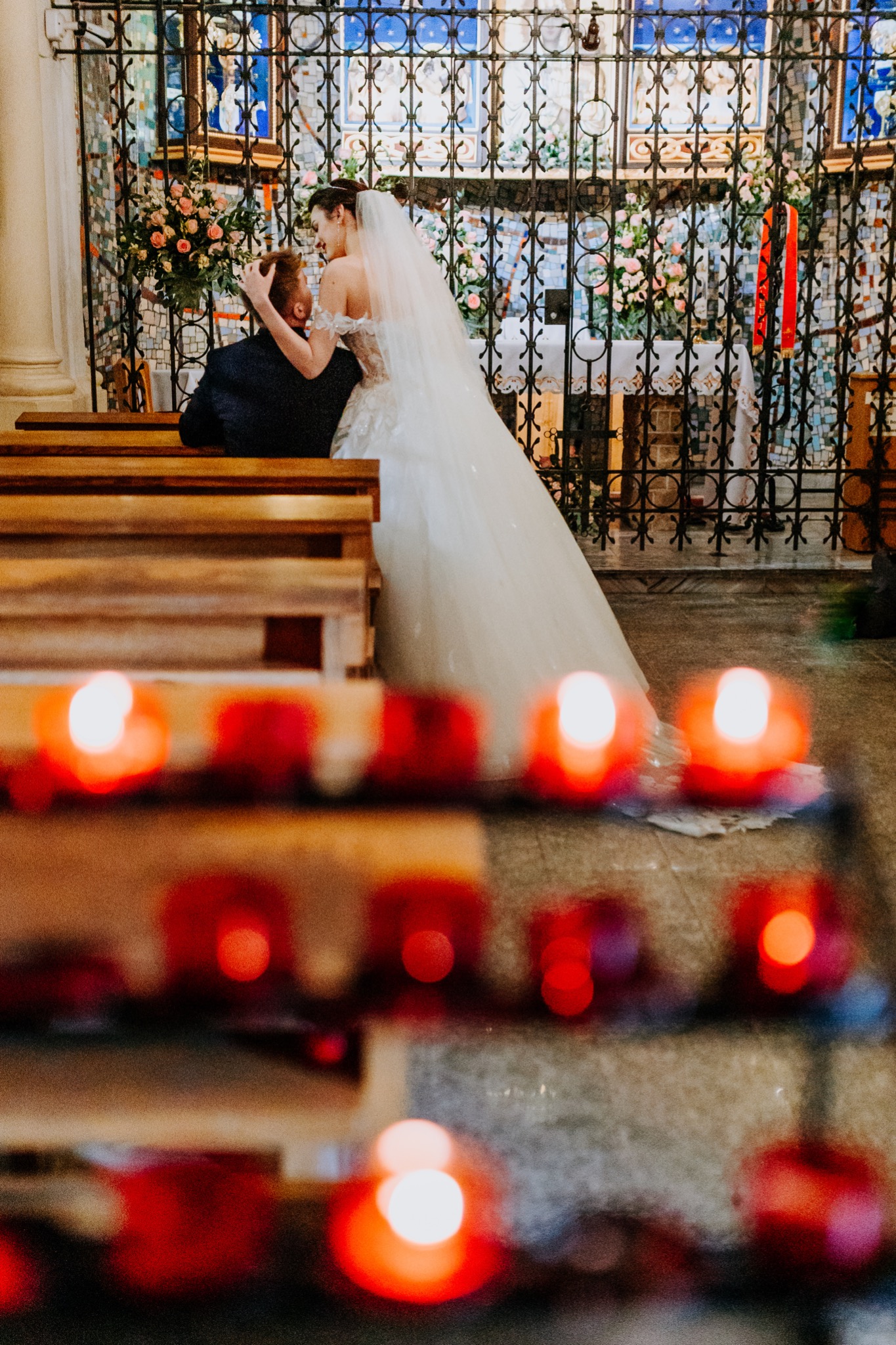 Para młoda w kościele, panna młoda w białej sukni i welonie obejmuje pana młodego na tle dekoracji kwiatowych i świec.