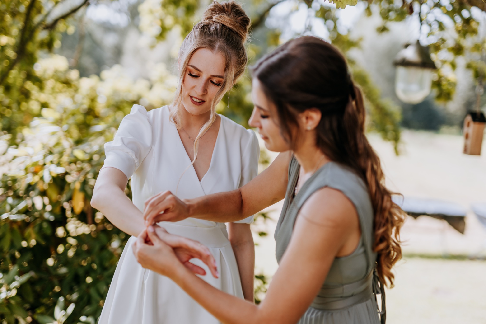 Panna młoda w białej sukni, której druhna zapina bransoletkę na nadgarstku w plenerze.