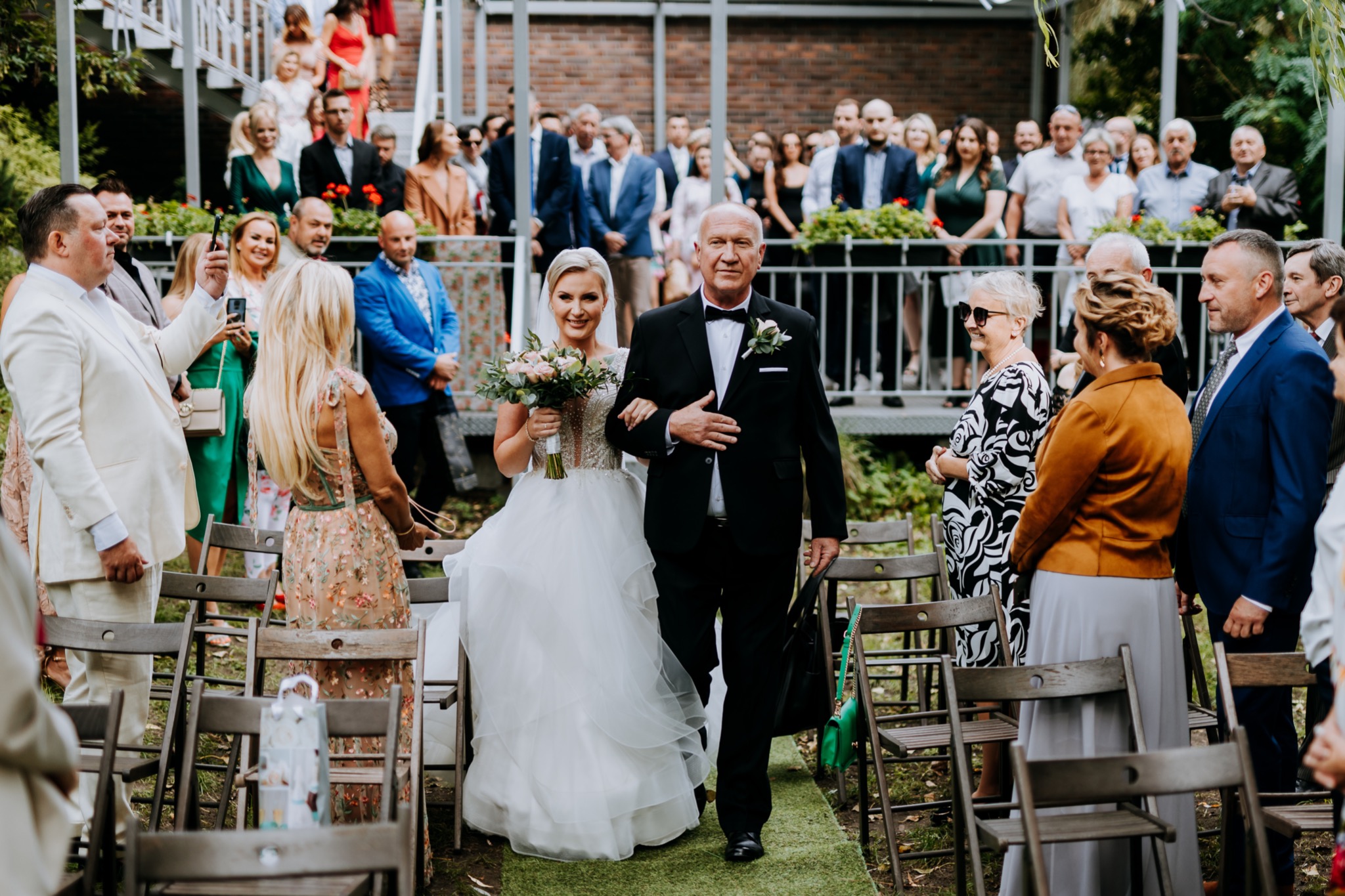 Panna młoda prowadzona przez ojca w dół ścieżki podczas ceremonii ślubnej na świeżym powietrzu, goście obserwują z boku i z balkonu.