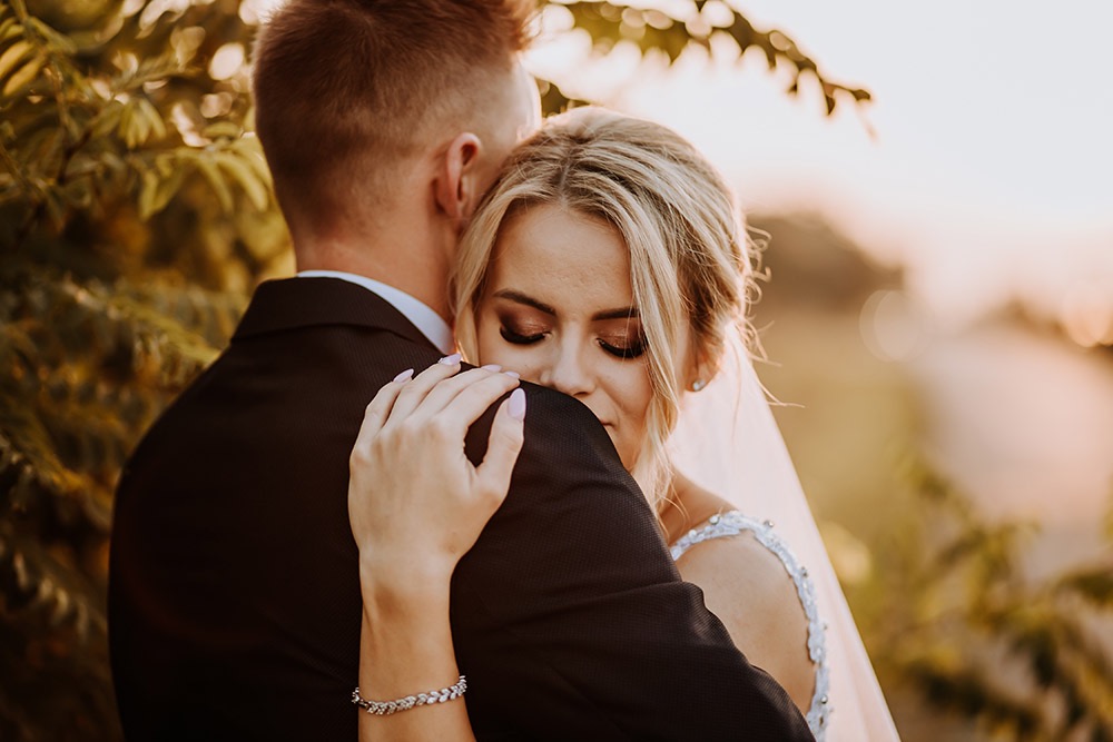 Panna młoda w białej sukni z koronkowymi ramiączkami, obejmująca pana młodego w ciemnym garniturze, subtelny makijaż i bransoletka na nadgarstku, ciepłe światło zachodzącego słońca.