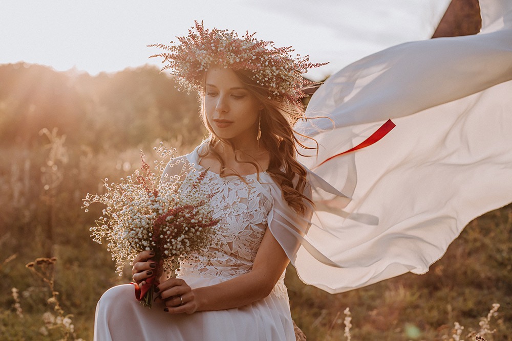 Panna młoda w białej sukni i wianku na głowie, trzymająca bukiet polnych kwiatów, z powiewającą tkaniną w tle, w ciepłym świetle zachodzącego słońca.