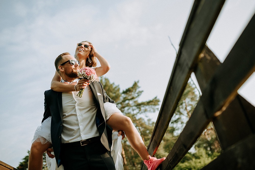 Uśmiechnięta panna młoda w okularach przeciwsłonecznych na barana u pana młodego, trzyma bukiet róż, różowe sneakersy na drewnianej konstrukcji, widok z dołu.