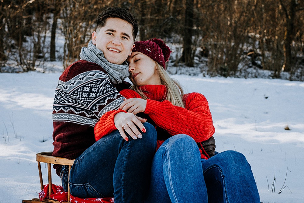 Uśmiechnięta para w zimowych ubraniach siedzi na sankach na śniegu; kobieta opiera głowę na ramieniu mężczyzny, w tle drzewa bez liści.