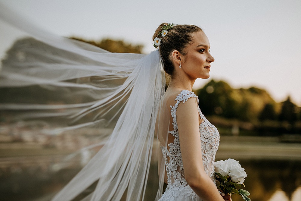 Panna młoda w koronkowej sukni, z wpiętymi kwiatami we włosach i welonem powiewającym na wietrze, profil na tle zieleni i wody.
