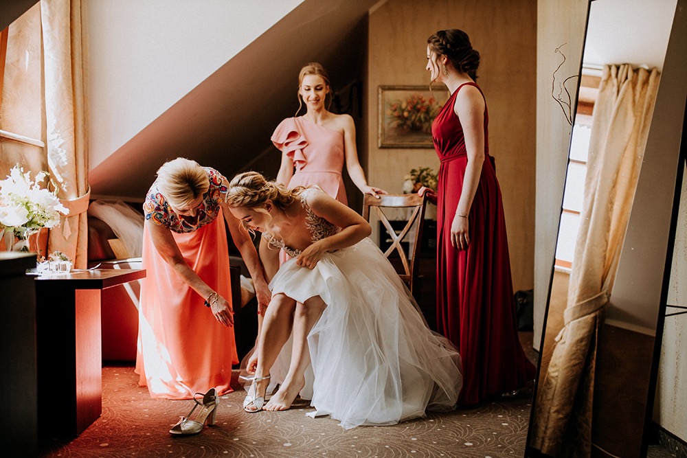 Panna młoda w białej sukni ślubnej zakłada srebrne buty z pomocą kobiety w pomarańczowej sukience, w tle dwie druhny w różowej i bordowej sukience, przygotowania ślubne w pokoju z lustrem i kwiatami.