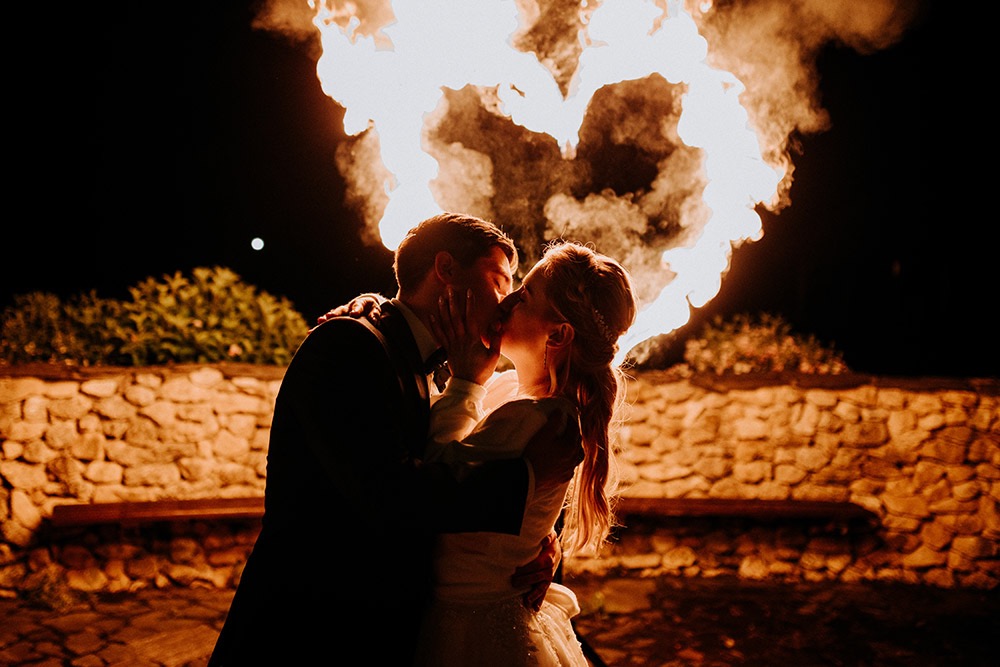 Pocałunek pary młodej na tle płonącego serca z ognia podczas nocnej ceremonii, kamienny murek i krzewy w tle.
